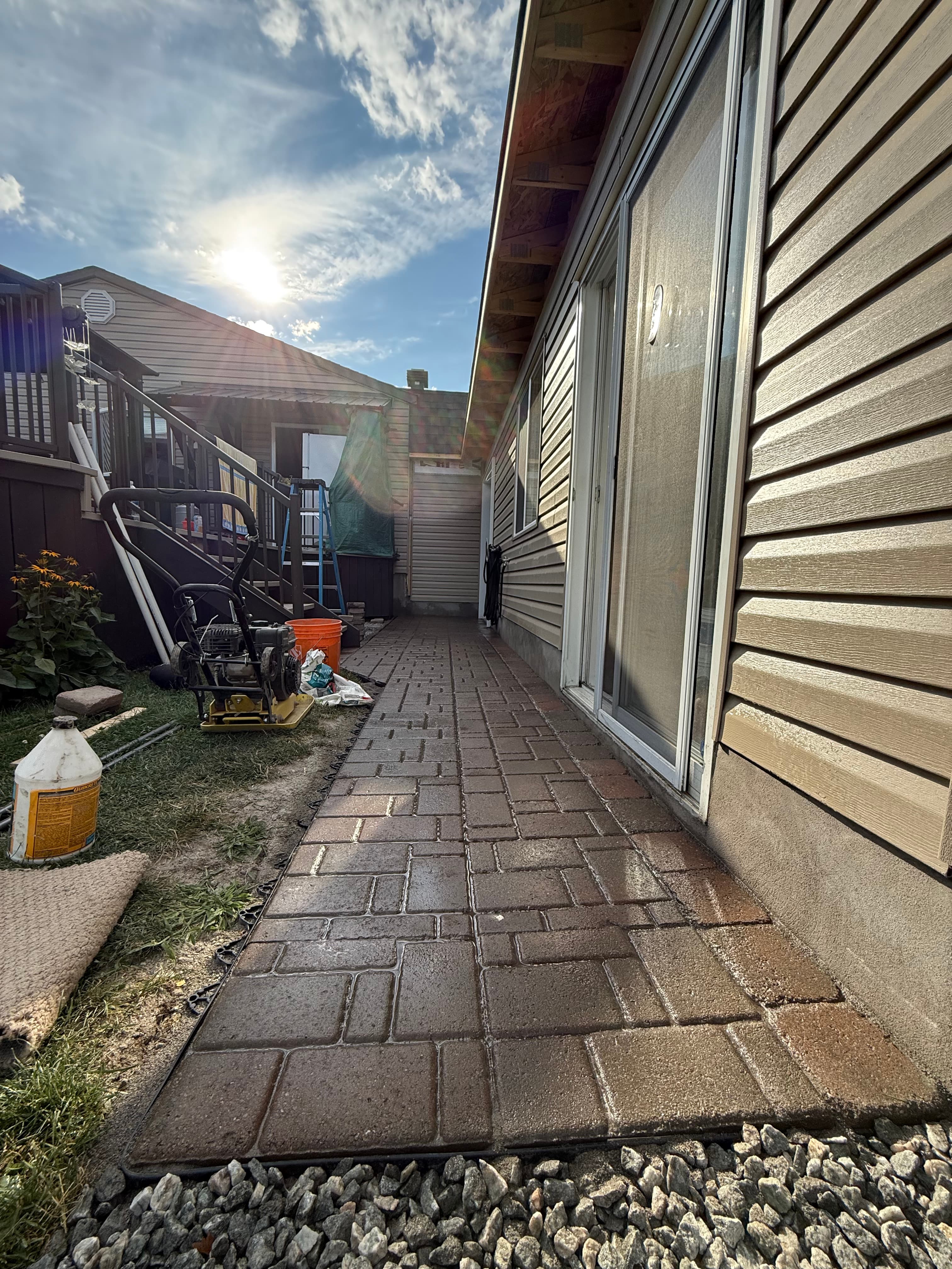 Wet brick paver walkway beside a beige house with construction equipment under bright sunlight.