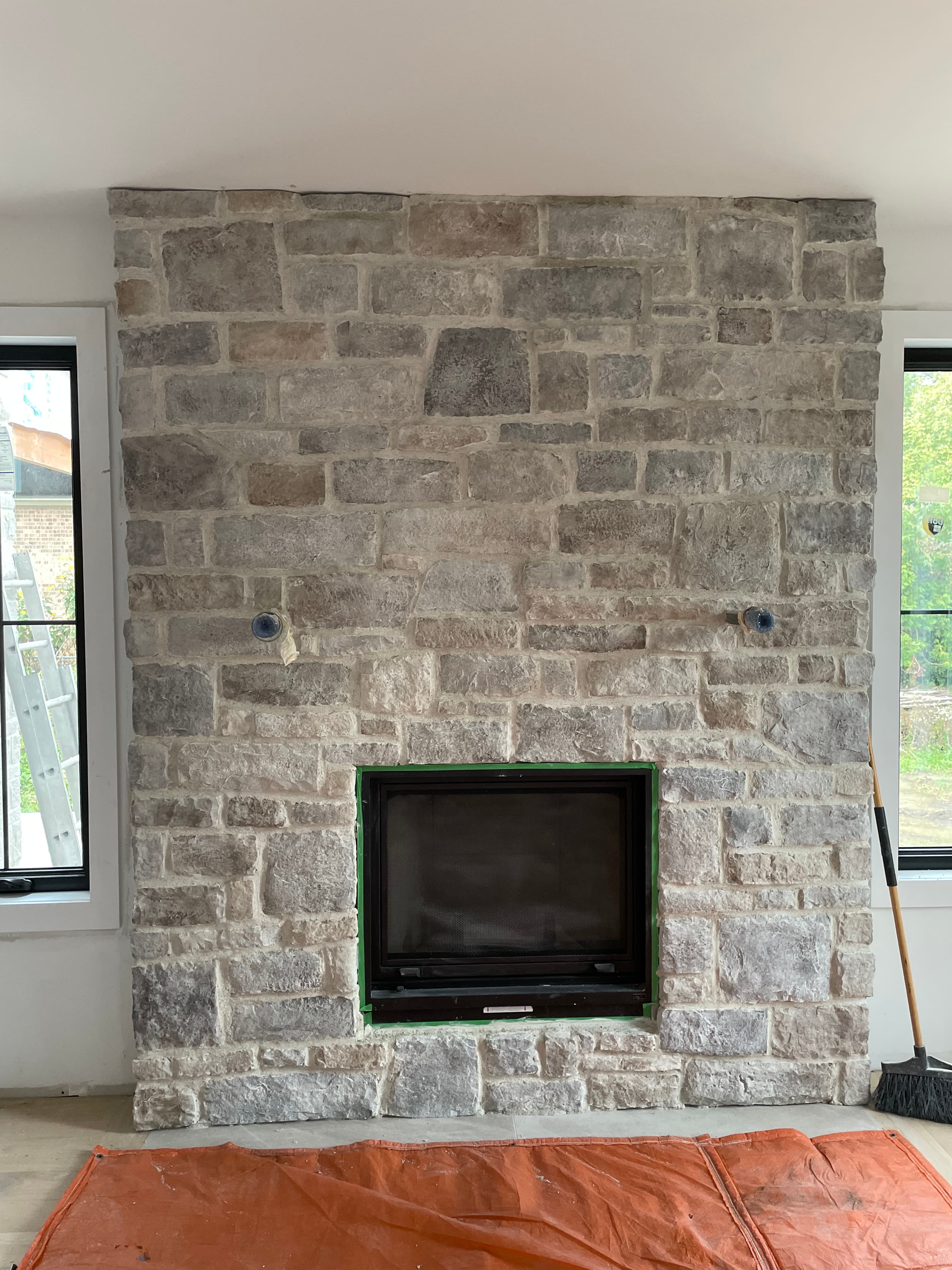 Light gray stone fireplace wall with a black insert under construction between two large windows.