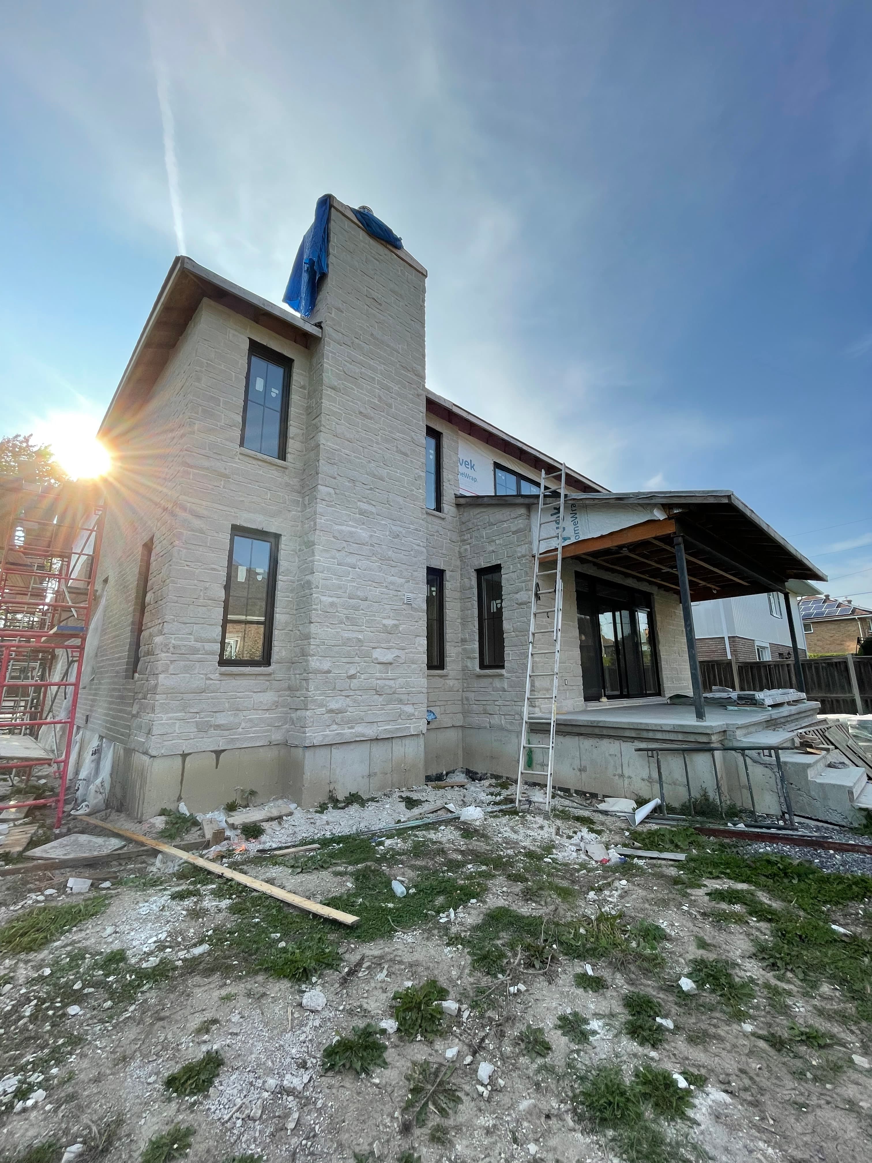 Modern stone house under construction with bright sun flare and scaffolding on a clear day.